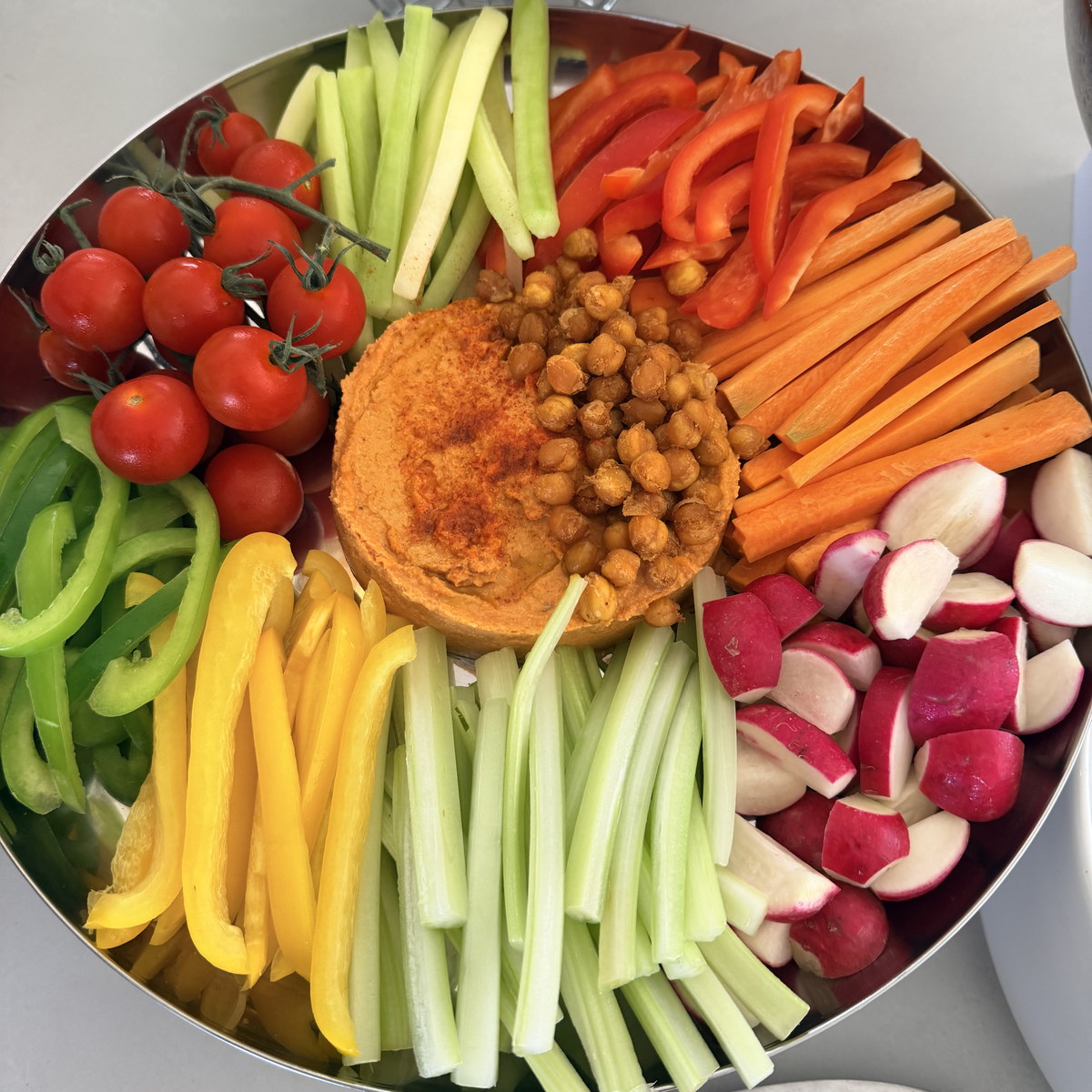A round platter of fresh vegetables around hummus and roasted chickpeas